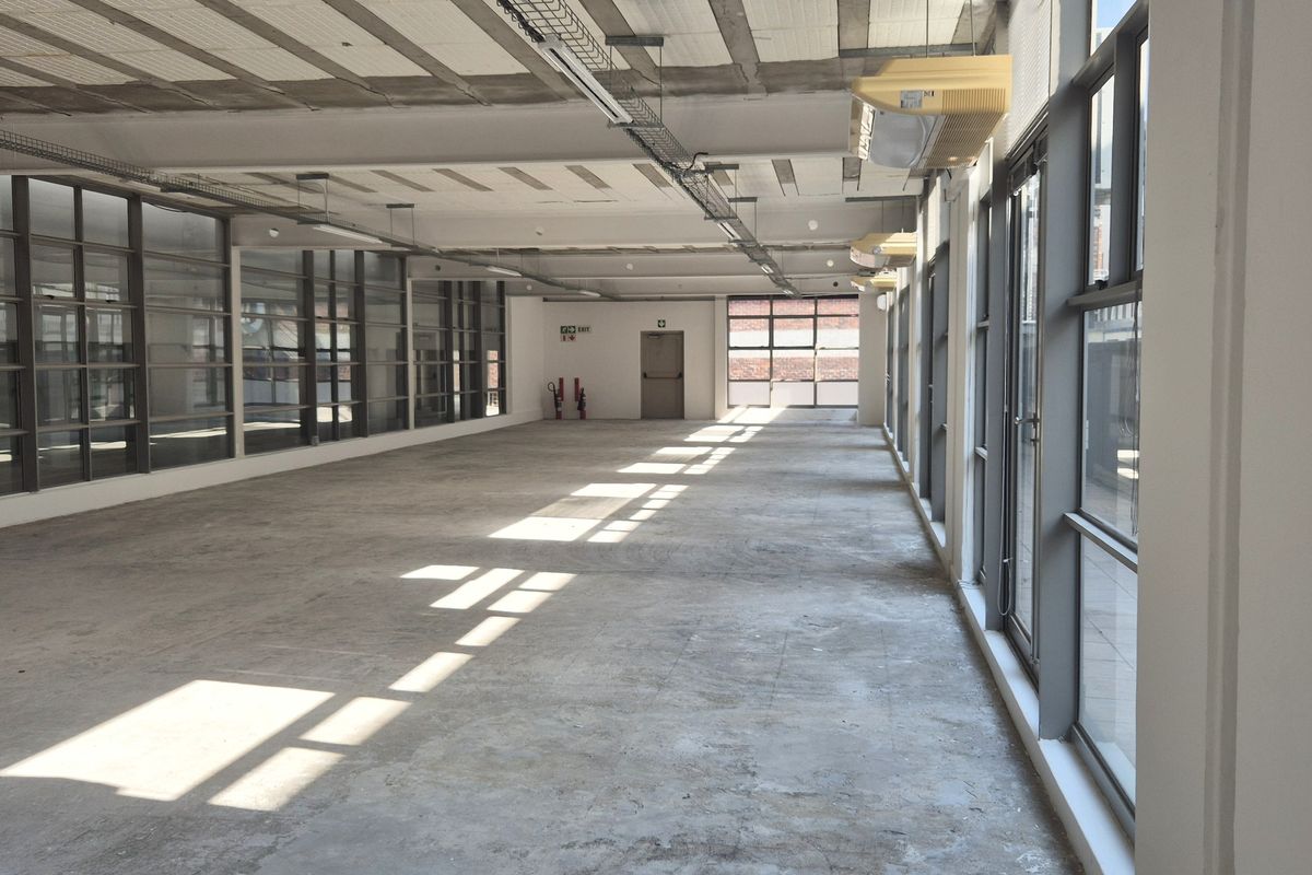 A large, open-plan office space featuring exposed concrete ceilings, extensive natural light from large windows, and industrial-style exposed cable trays.
