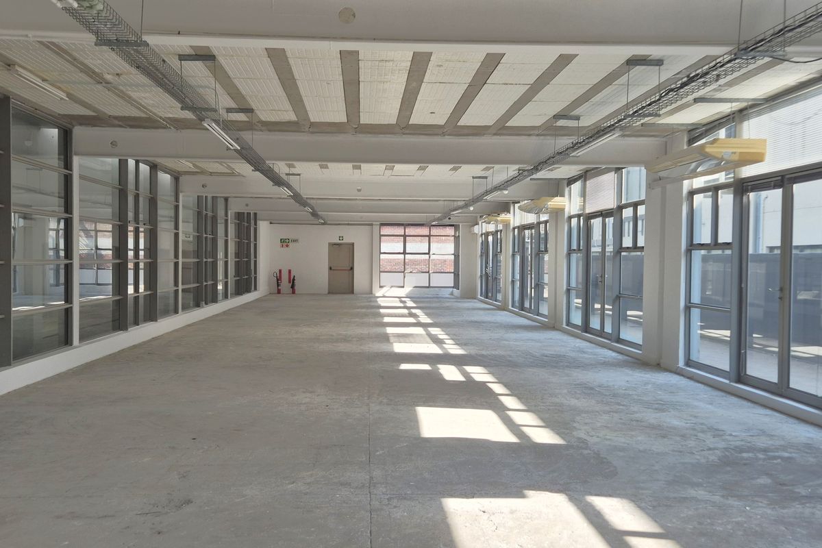 Spacious open-plan office area featuring industrial-style exposed ceilings, concrete flooring, and extensive natural light through large aluminium-framed windows.