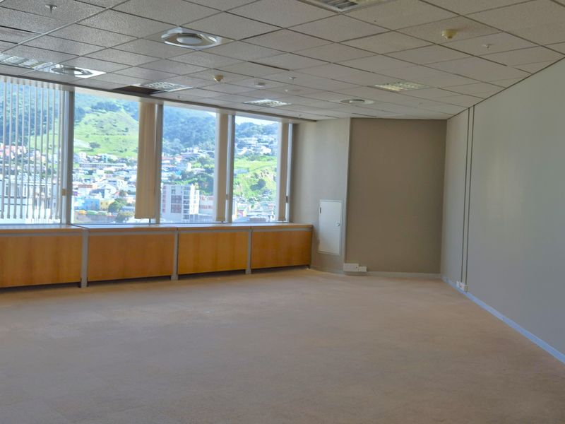A spacious, empty office area featuring large windows with city views, a suspended ceiling, and neutral carpeted flooring.
