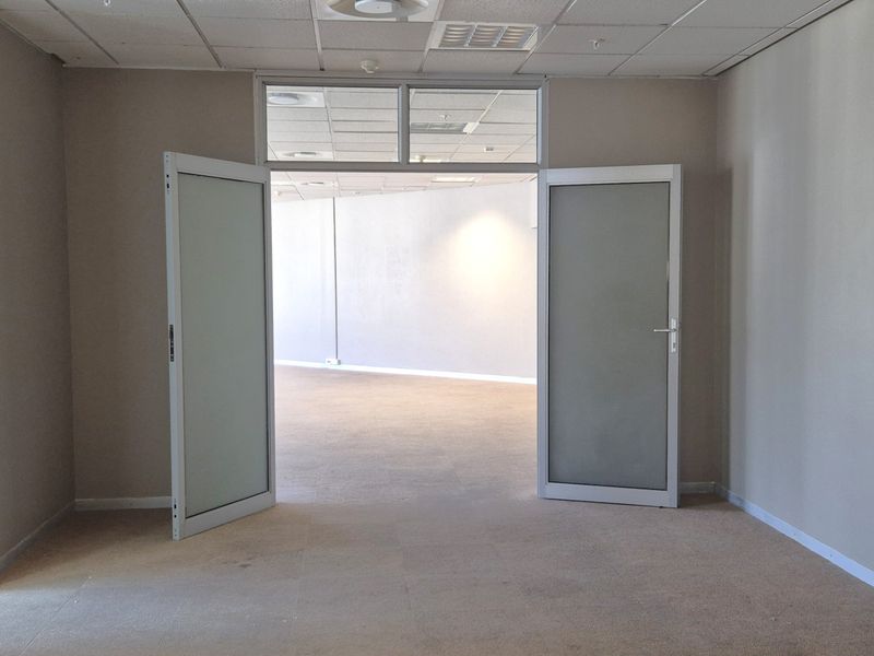 An empty, well-lit office space featuring neutral carpeted flooring, a suspended ceiling, and frosted glass double doors leading to an adjacent area.