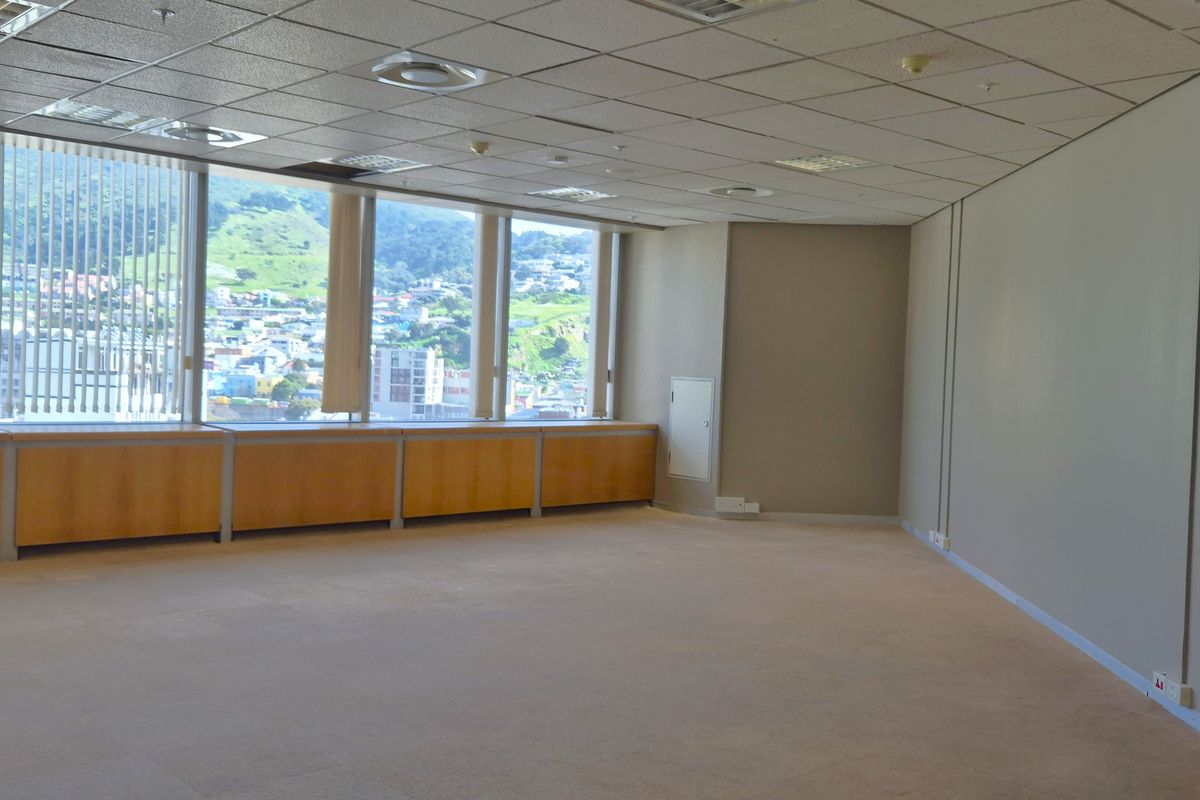A spacious, empty office area featuring large windows with city views, a suspended ceiling, and neutral carpeted flooring.