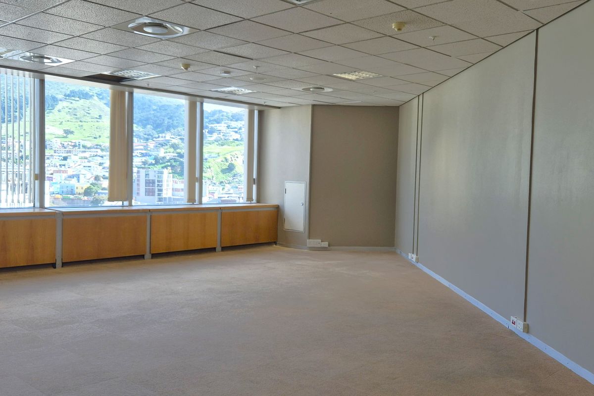 A spacious, empty office area featuring large windows with city views, suspended ceilings, recessed lighting, and built-in wooden cabinetry along the window perimeter.