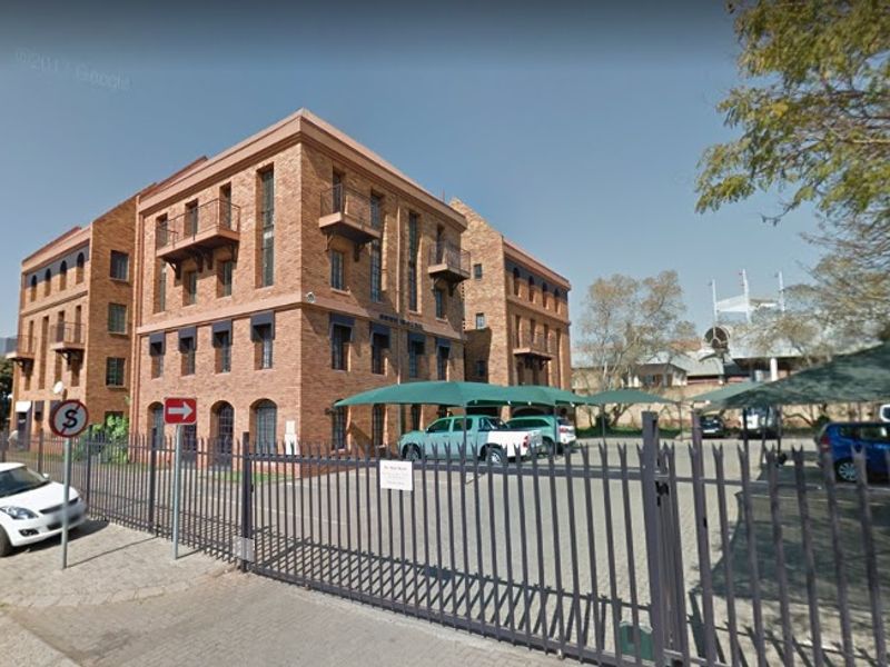 A multi-story brick office building featuring balconies, secure perimeter fencing, and covered parking bays within a paved courtyard area.