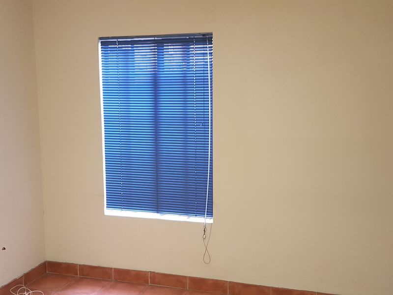 A simple, empty office space featuring terracotta floor tiles, neutral walls, and blue venetian blinds.