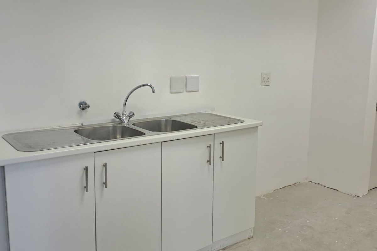 A functional kitchenette area featuring a stainless steel double sink, chrome faucet, and white cabinetry, set within an unfinished office space.