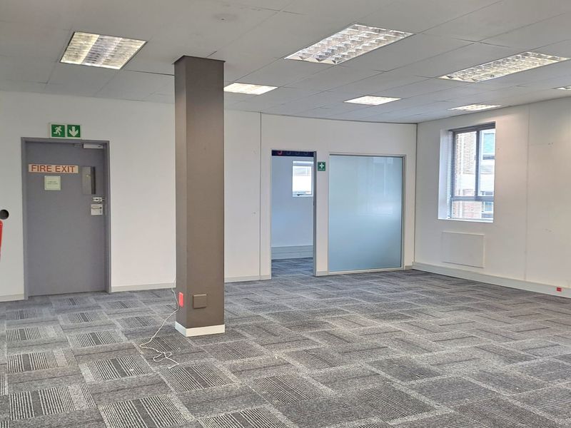 A bright, open-plan office space featuring carpet tiles, suspended ceilings with recessed lighting, and a functional fire exit door.