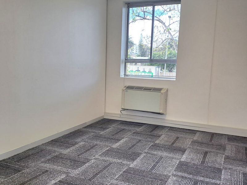A vacant office space featuring grey patterned carpet tiles, a suspended ceiling, and a wall-mounted air conditioning unit.