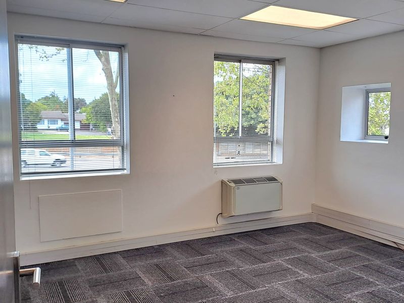 A bright, empty office space featuring carpet tiles, suspended ceiling with recessed lighting, and a wall-mounted air conditioning unit.