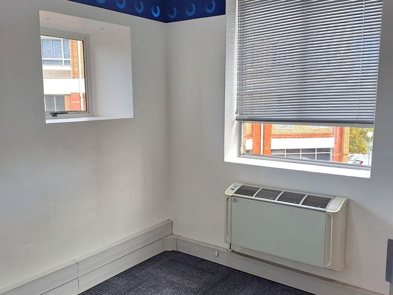 A vacant office space featuring carpet tiles, a wall-mounted HVAC unit, and dual windows with venetian blinds.