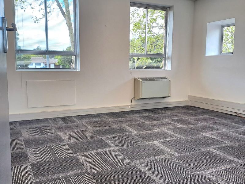 A bright, empty office space featuring carpet tiles, a suspended ceiling with recessed lighting, and a wall-mounted HVAC unit.