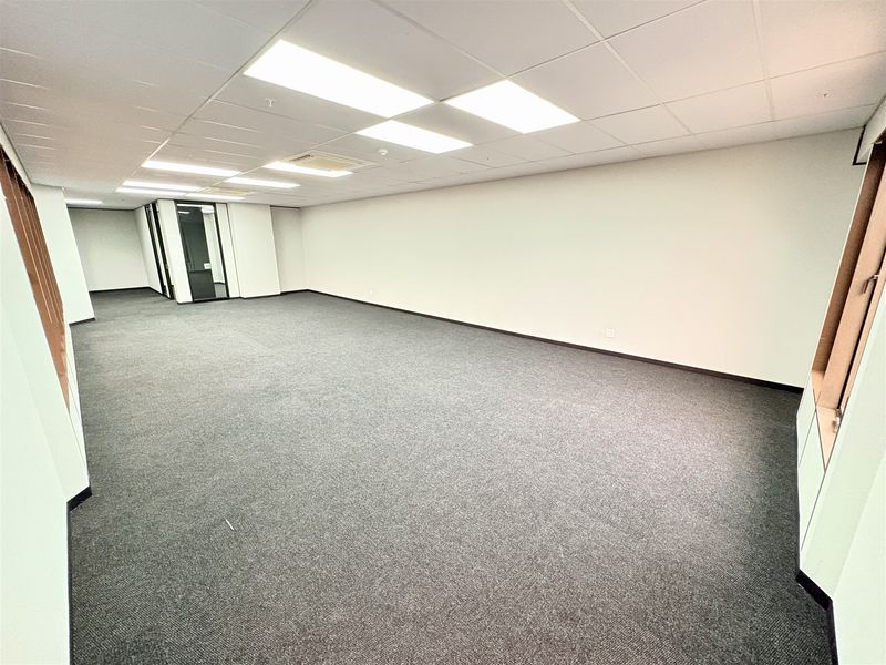 A spacious, empty open-plan office area featuring neutral carpeting, a suspended ceiling with recessed lighting, and a modern glass partition entrance.