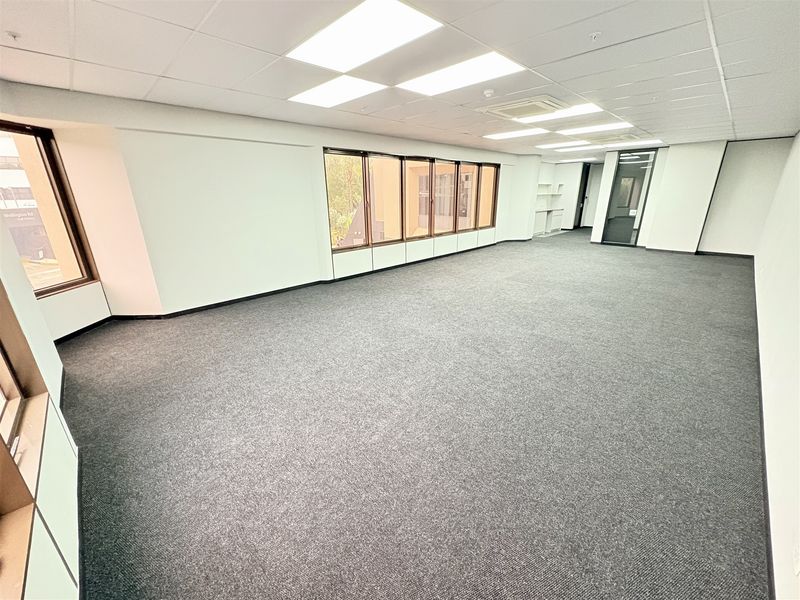 A spacious, empty open-plan office area featuring neutral carpet flooring, recessed ceiling lighting, and large windows providing ample natural light.