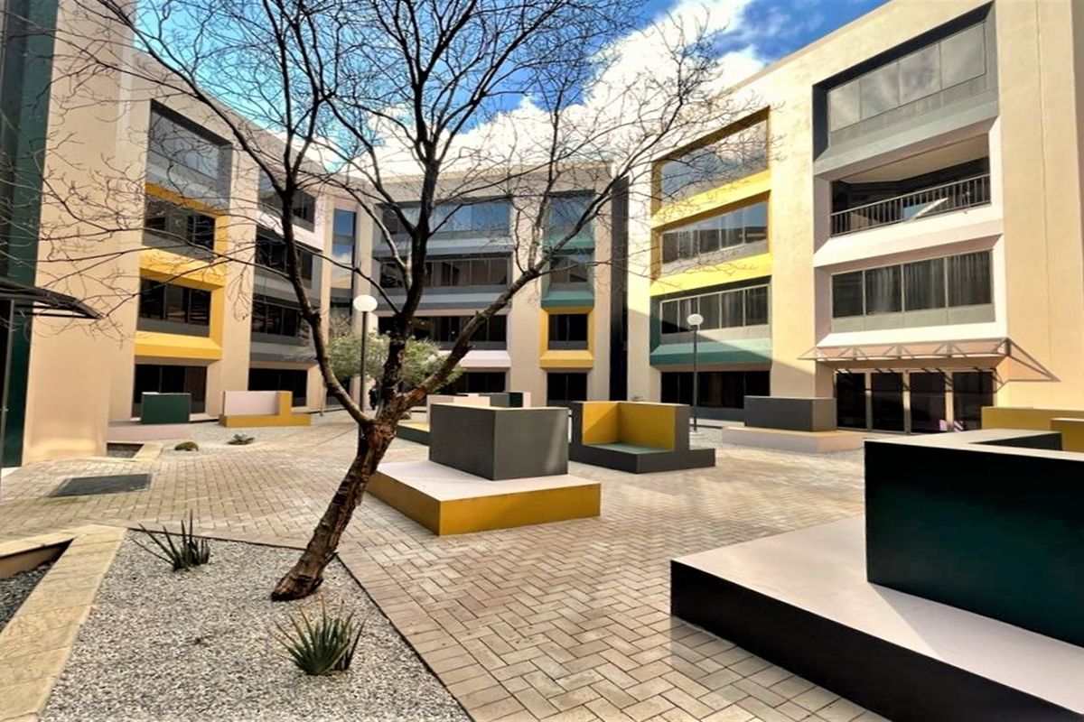 A modern office complex courtyard featuring paved walkways, built-in concrete seating areas, and multi-story building facades with large glass windows.