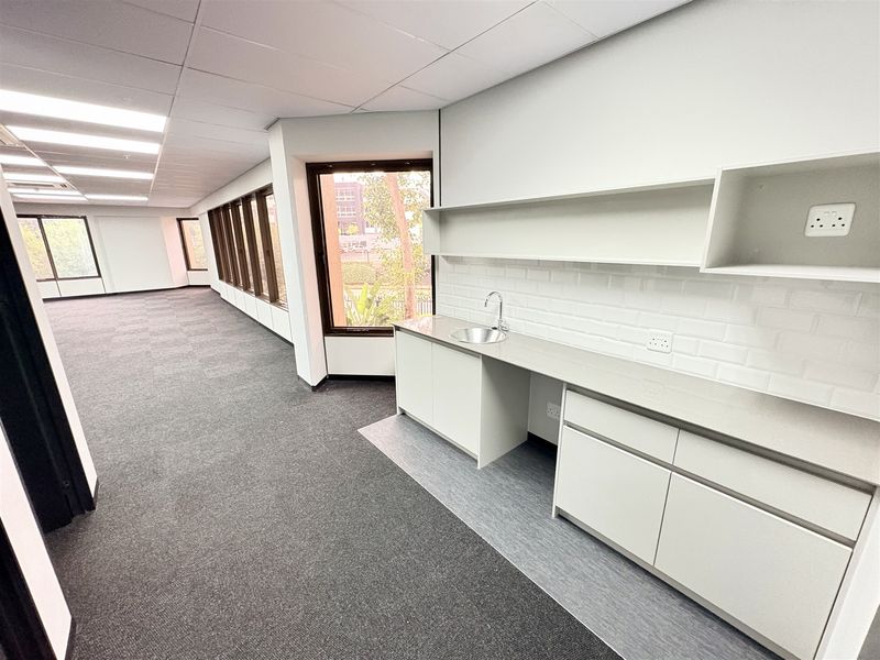 Spacious open-plan office area featuring a modern built-in kitchenette, neutral finishes, carpeted flooring, and large windows providing ample natural light.