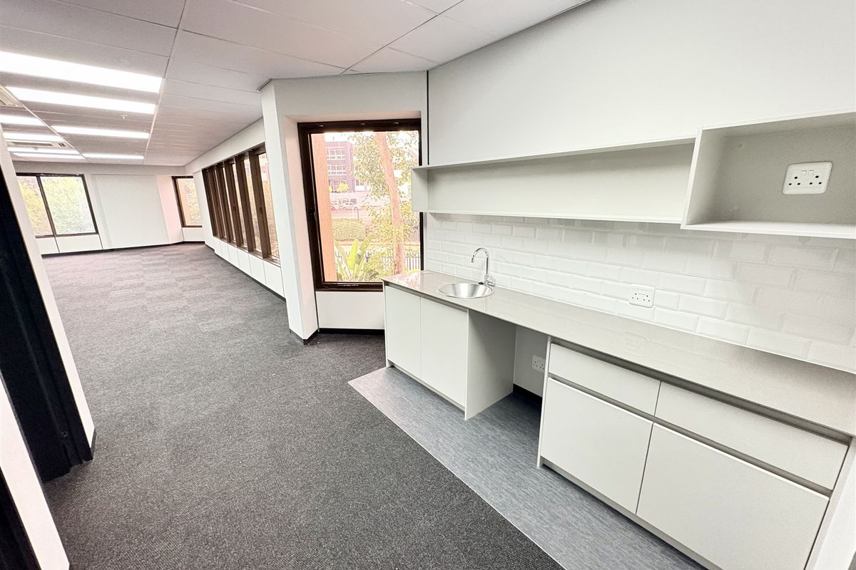 Spacious open-plan office area featuring a modern built-in kitchenette, neutral finishes, carpeted flooring, and large windows providing ample natural light.