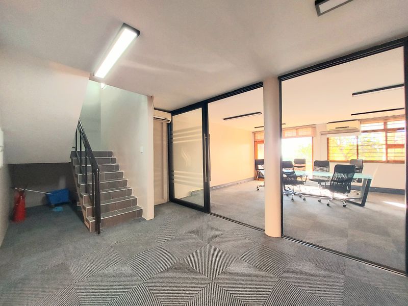 A professional office space featuring a glass-partitioned meeting room, carpeted flooring, and an internal staircase leading to another level.