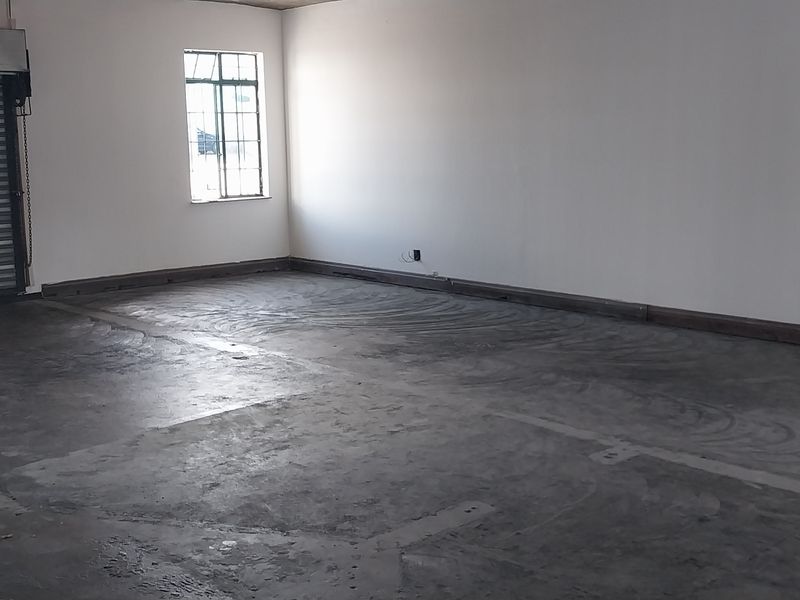 An open-plan industrial utility space featuring a roller shutter door, concrete flooring, and a steel-framed window, suitable for storage or workshop use.