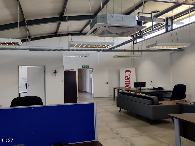 Open-plan industrial office space featuring exposed steel roof trusses, ceiling-mounted air conditioning, and tiled flooring with ample natural light.