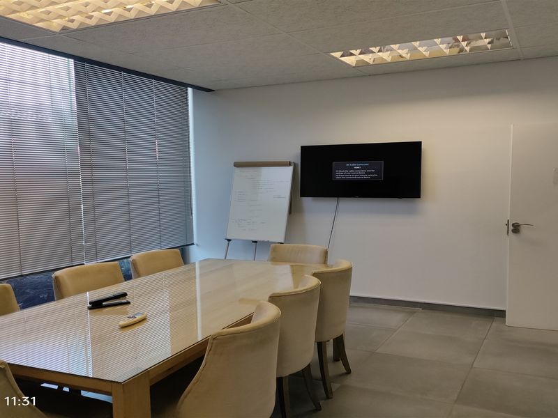 A functional office meeting room featuring tiled flooring, a suspended ceiling with recessed lighting, and a wall-mounted television for presentations.