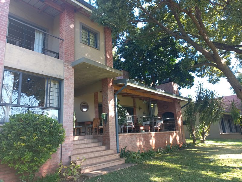 A rear view of a multi-story brick farmhouse featuring a spacious covered patio with a built-in braai and an upper-level balcony.