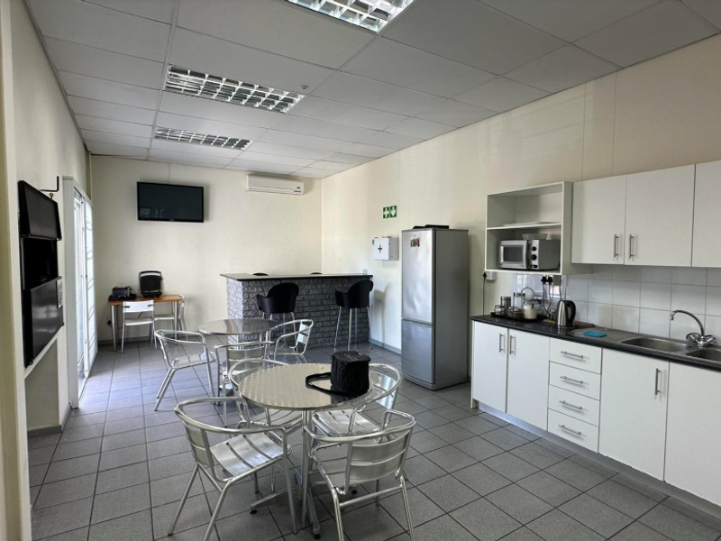 Open-plan industrial breakroom featuring a kitchenette with white cabinetry, tiled flooring, suspended ceiling, and a brick-faced breakfast bar area.
