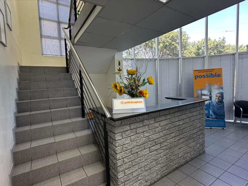 A bright industrial reception area featuring a brick-faced reception desk, tiled flooring, a staircase with metal railings, and large floor-to-ceiling windows.