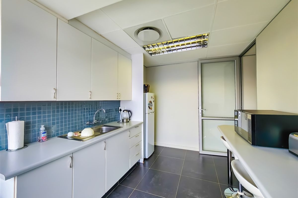A functional office kitchenette featuring built-in cabinetry, a stainless steel sink, tiled backsplash, and a breakfast bar area with overhead lighting.