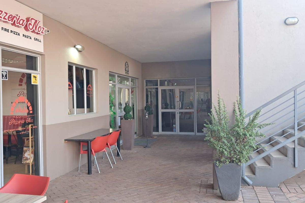 Covered outdoor patio area at a retail pizzeria, featuring paved flooring, access to an external staircase, and glass storefront entrance doors.