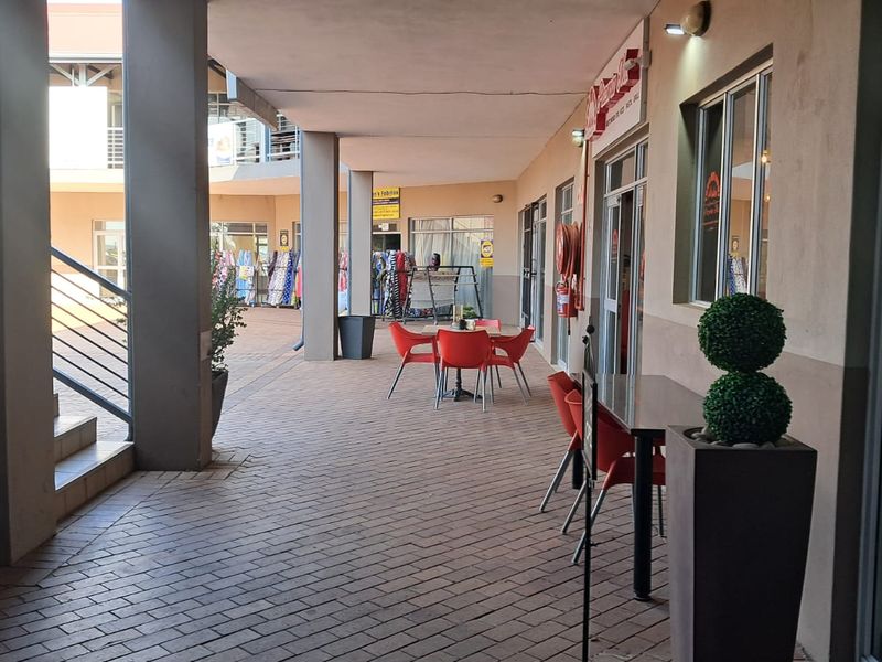 A covered outdoor walkway and patio area in a retail complex, featuring paved flooring, concrete support pillars, and glass storefront access.