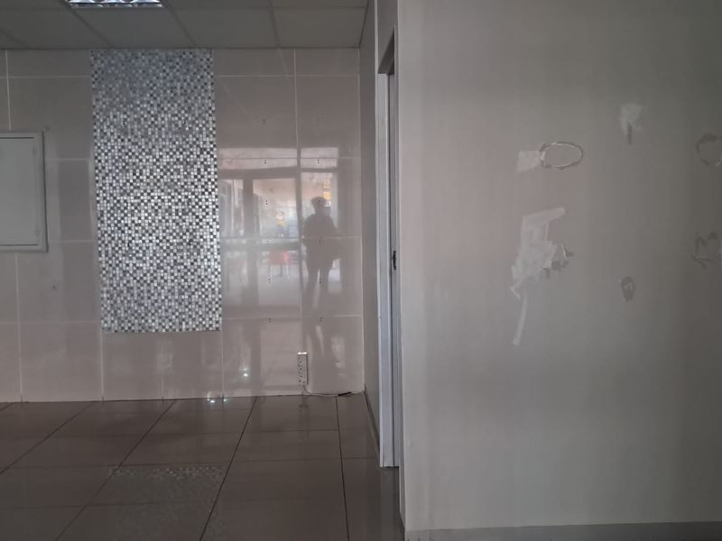 An empty retail space featuring tiled flooring, a suspended ceiling, and a decorative mosaic wall accent, currently requiring minor wall repairs.