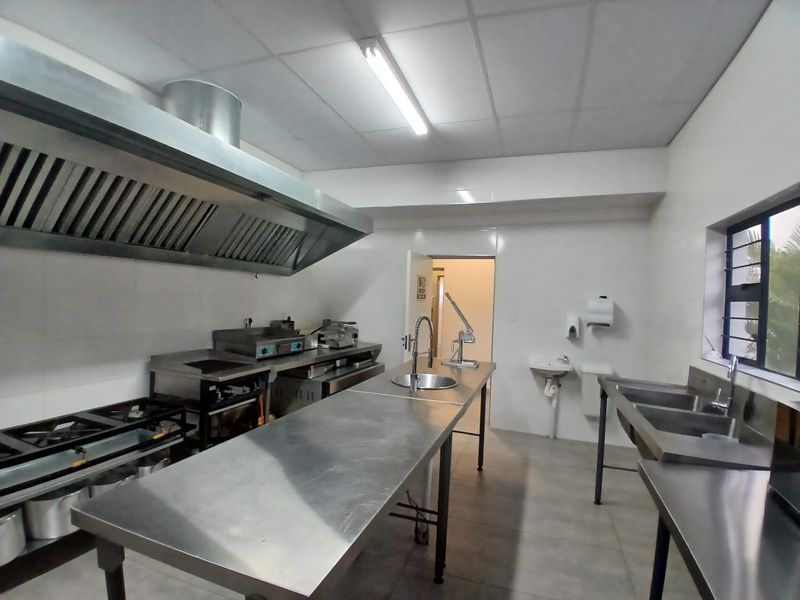 A functional commercial kitchen featuring stainless steel prep surfaces, industrial gas hobs, a large extractor hood, and a double sink setup.