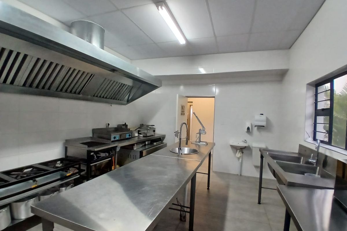 A functional commercial kitchen featuring stainless steel prep surfaces, industrial gas hobs, a large extractor hood, and a double sink setup.