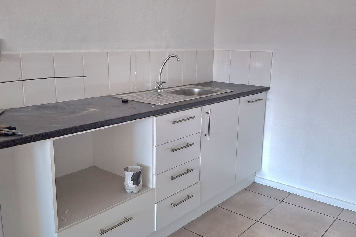 A functional kitchenette area featuring white cabinetry, a stainless steel sink, dark laminate countertop, and tiled splashback.