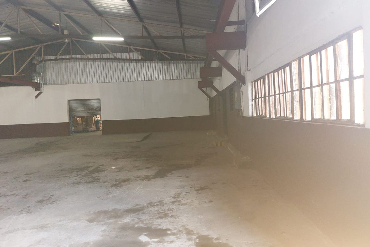 Spacious industrial warehouse area featuring high exposed steel truss ceilings, concrete flooring, and large side windows providing natural light.