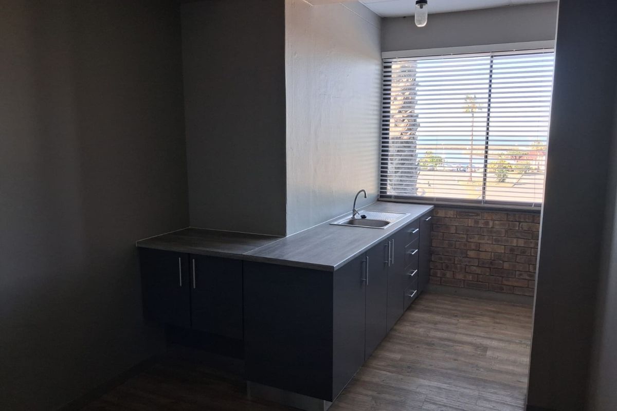 A compact office kitchenette featuring modern dark cabinetry, a stainless steel sink, and wood-look flooring with natural light from a large window.