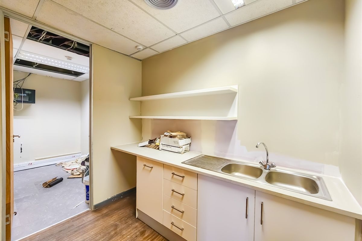 A small office kitchenette featuring built-in cabinetry, a double stainless steel sink, and open shelving, with an adjacent room showing signs of ongoing maintenance.