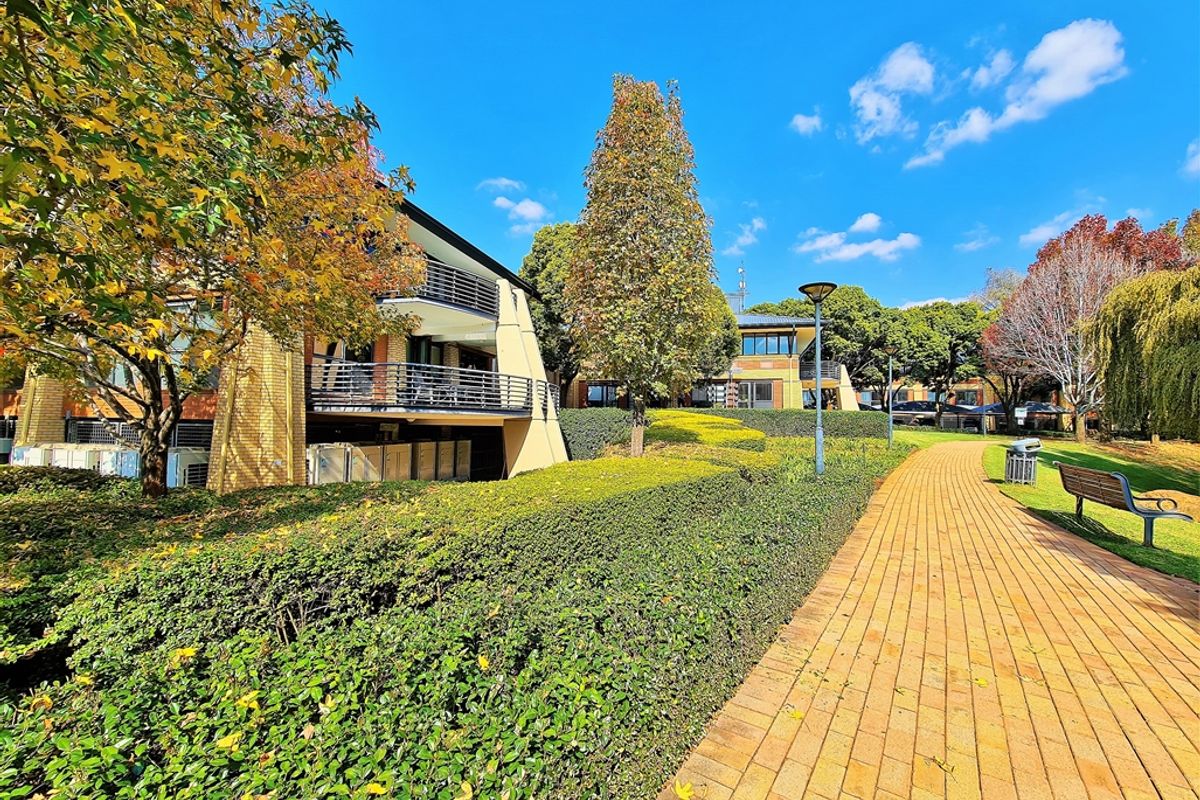 A well-maintained office park exterior featuring paved walkways, manicured hedges, mature trees, and modern multi-story office buildings with balconies.