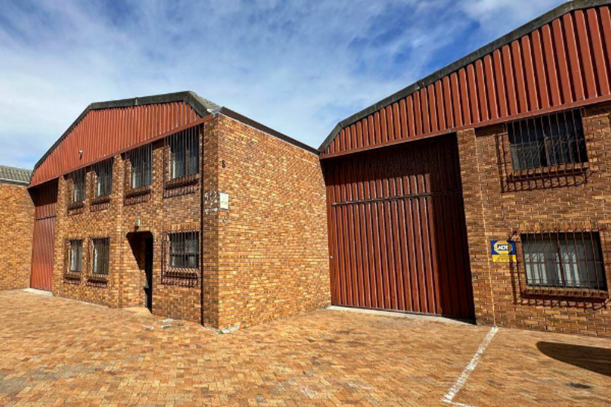 Industrial property exterior featuring face brick construction, corrugated metal roofing, security window bars, and a large roller shutter door with paved frontage.