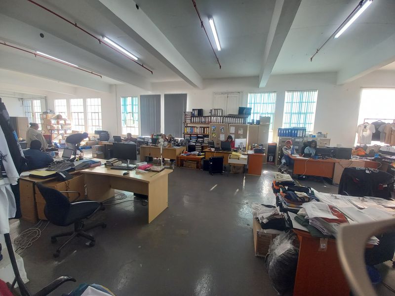 An open-plan industrial office space featuring high ceilings, exposed structural beams, fluorescent lighting, and large multi-pane windows providing natural light.