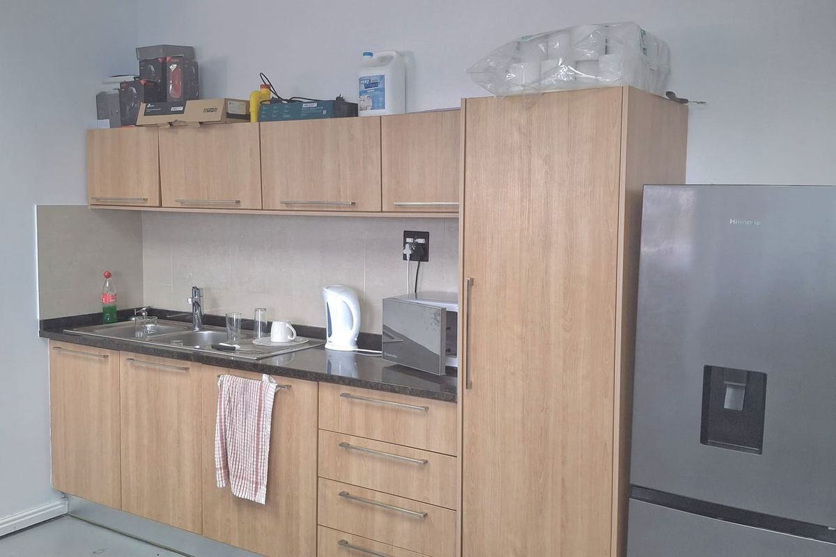 A functional office kitchenette featuring light wood cabinetry, a dark countertop, integrated sink, and a tall pantry cupboard.