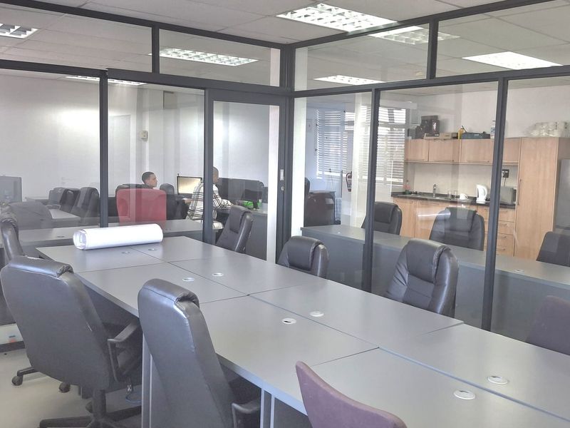 A professional office space featuring glass-partitioned meeting rooms, a central conference table, and a small kitchenette area with built-in cabinetry.
