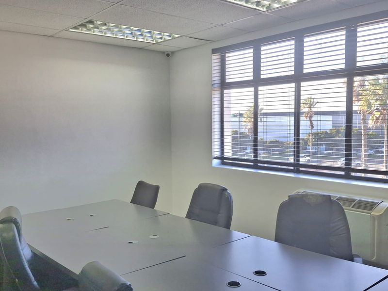 A functional office meeting room featuring recessed ceiling lighting, large windows with blinds, and a wall-mounted HVAC unit.