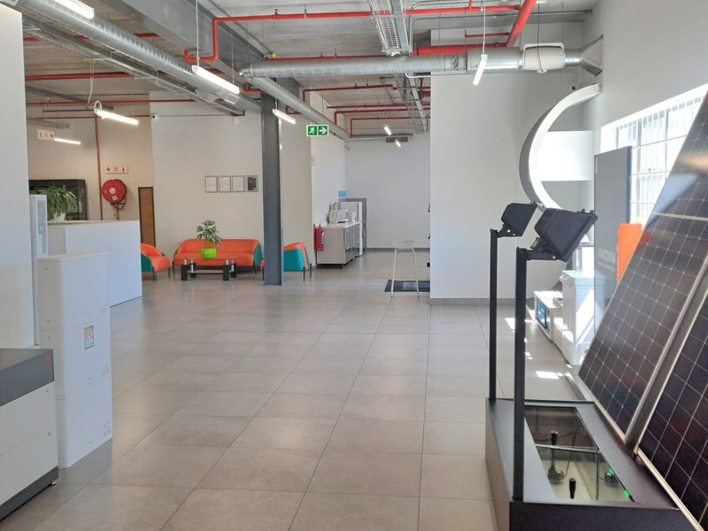 Spacious industrial open-plan area featuring exposed ceiling infrastructure, fire suppression systems, tiled flooring, and large windows, suitable for showroom or office use.