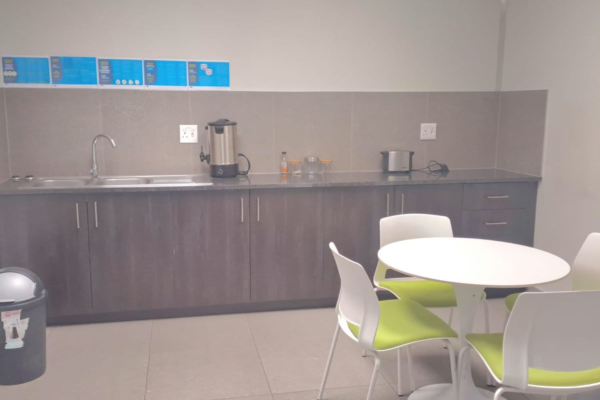 A functional kitchenette area featuring dark cabinetry, a stainless steel sink, tiled backsplash, and tiled flooring, suitable for office or industrial staff use.