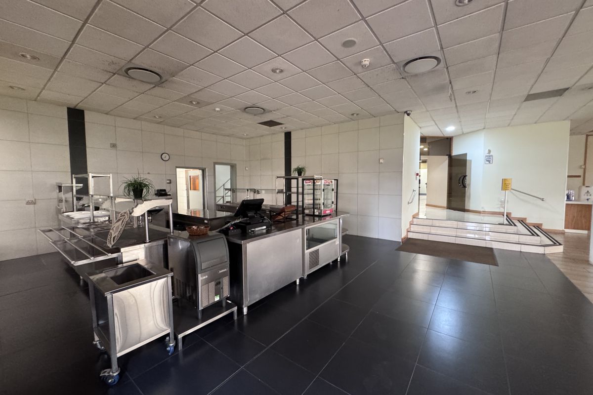 Industrial kitchen area featuring stainless steel preparation counters, tiled flooring, and a suspended ceiling with recessed lighting, leading to an elevated hallway.