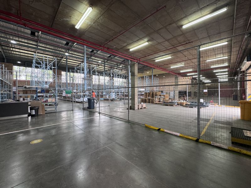 Spacious industrial warehouse area featuring concrete flooring, exposed ceiling with fire sprinkler system, and metal mesh partitioning for secure storage zones.