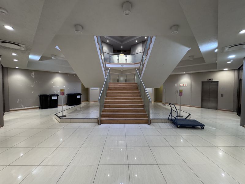 Spacious industrial building entrance lobby featuring a central staircase with glass balustrades, tiled flooring, and elevator access.