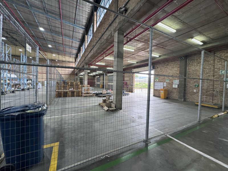 A spacious industrial warehouse area featuring exposed brick walls, concrete flooring, high-bay ceilings with fire sprinklers, and secure wire mesh partitioning.