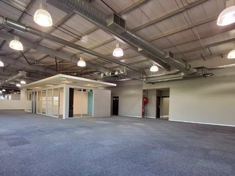 Spacious open-plan office area featuring industrial-style exposed ductwork, suspended lighting, carpeted flooring, and a central glass-enclosed meeting room or private office suite.