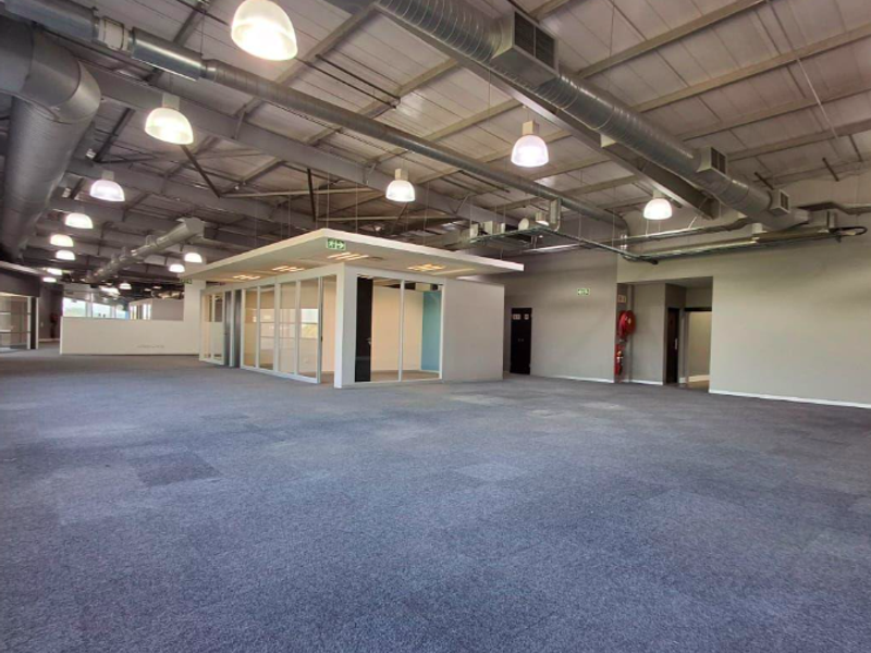 Spacious open-plan office area featuring exposed industrial ductwork, modern glass-partitioned meeting rooms, and durable carpet tiles throughout.